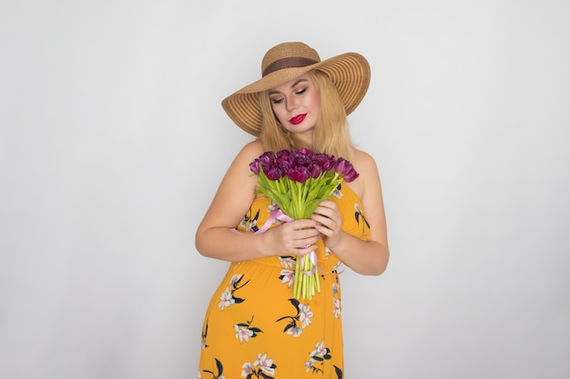 Mooie blonde vrouw in gele bloemen jurk en strooien hoed met boeket paarse tulpen