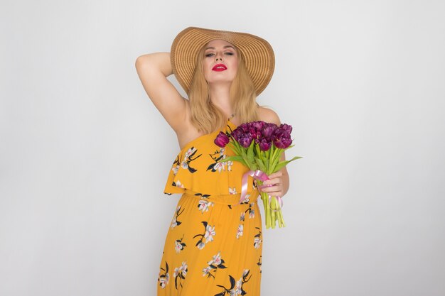 Mooie blonde vrouw in gele bloemen jurk en strooien hoed met boeket paarse tulpen