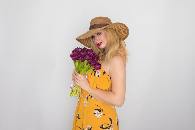 Mooie blonde vrouw in gele bloemen jurk en strooien hoed met boeket paarse tulpen