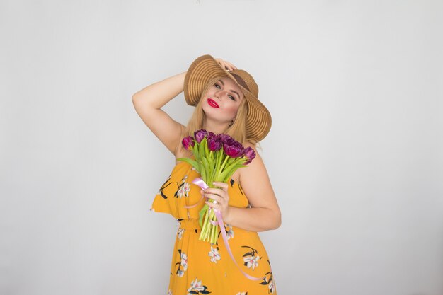 Mooie blonde vrouw in gele bloemen jurk en strooien hoed met boeket paarse tulpen