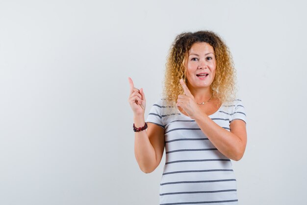 Mooie blonde vrouw in gestreept t-shirt wijzend naar de linkerbovenhoek en er vrolijk uitziend, vooraanzicht.