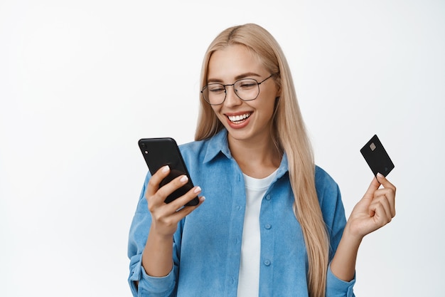 Mooie blonde vrouw in glazen met creditcard, met behulp van mobiele telefoon app en glimlachen, volgorde in toepassing, staande in casual kleding op wit.