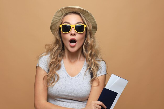 Mooie blonde vrouw in zonnebril poseren met paspoort met kaartjes over beige achtergrond.