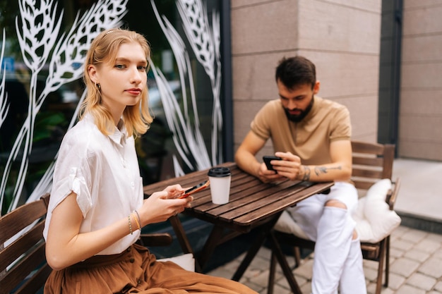 Mooie blonde vrouw kijken camera bebaarde getatoeëerde man met behulp van smartphones zittend aan tafel met koffie in straatcafé op zomerdag