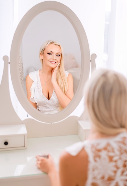 Mooie blonde vrouw kijkt in de spiegel en past crème op haar gezicht toe.