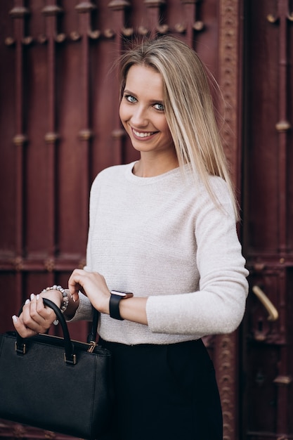 Mooie blonde vrouw kijkt naar de tijd op horloges, wandelen op straat. Portret van stijlvolle glimlachende zakenvrouw in donkere casual broek en romige trui. Mode concept. Vrouwelijke zakelijke stijl.