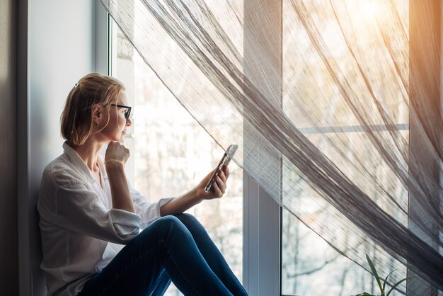 Mooie blonde vrouw met blote voeten in een bril, jeans en wit overhemd zittend op de vensterbank in zonlicht, smartphone in haar hand houden. Ontspanning in een gezellig interieur. Zijaanzicht, close-up.