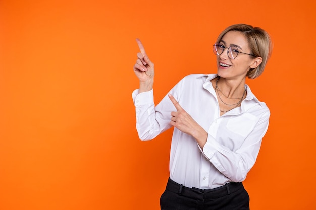 Mooie blonde vrouw met een wit shirt in officiële stijl wijst naar een kopieruimte voor reclame geïsoleerd op oranje achtergrond