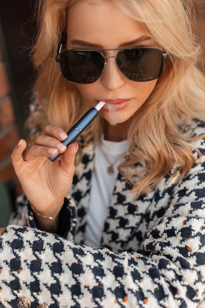 Mooie blonde vrouw model in modieuze uitloper met zonnebril rookt een e-sigaret op straat