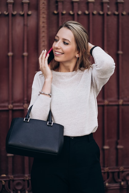 Mooie blonde vrouw praten over telefoon lopen op straat. Portret van stijlvolle glimlachende zakenvrouw in donkere casual broek en romige trui. Mode concept. Vrouwelijke zakelijke stijl. Hoge resolutie.