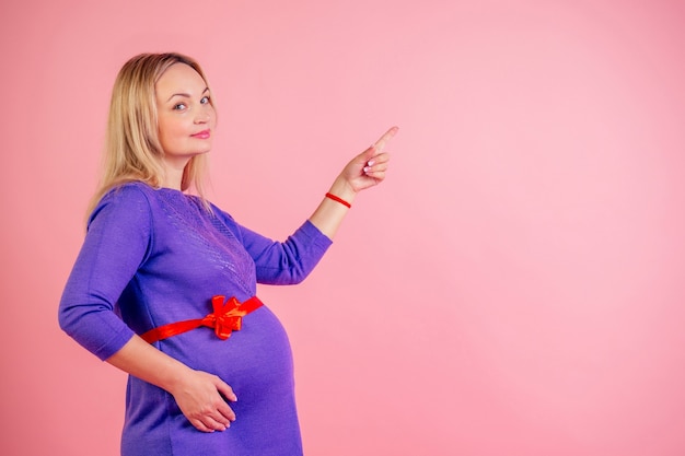 Mooie blonde zwangere vrouw in een jurk met rode satijnen strik op de buik droom van de babybuil en wijs met de vinger omhoog in de studio op een roze achtergrond copyspace.