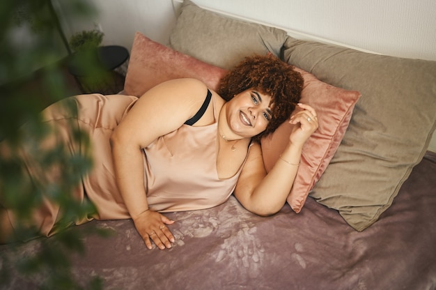 Mooie bochtige plus size Afrikaanse zwarte vrouw afro haar liggend op bed in zijden poederroze jurk gezellig