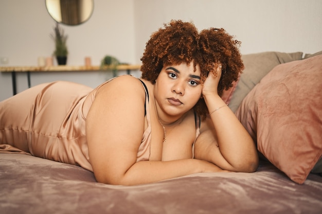 Mooie bochtige plus size Afrikaanse zwarte vrouw afro haar liggend op bed in zijden poederroze jurk gezellig