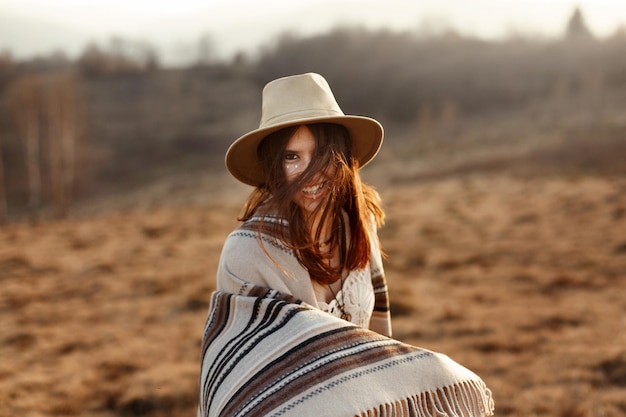 Foto mooie boho vrouw hipster glimlachen en plezier hebben bij zonsondergang in bergen met hoed en poncho