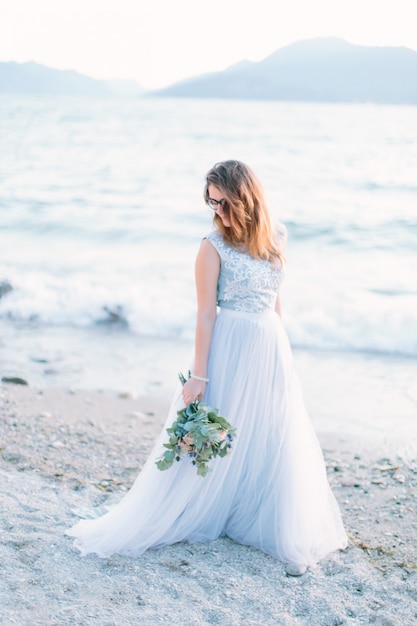 Mooie bruid in blauwe elegante jurk houdt een boeket bloemen lopen op de oevers van het gardameer. sirmione, italië