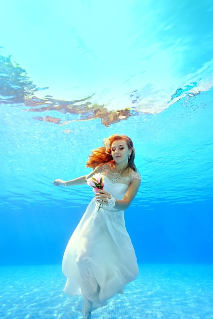 Mooie bruid in een witte jurk zwemt onder water met een bloem in zijn hand