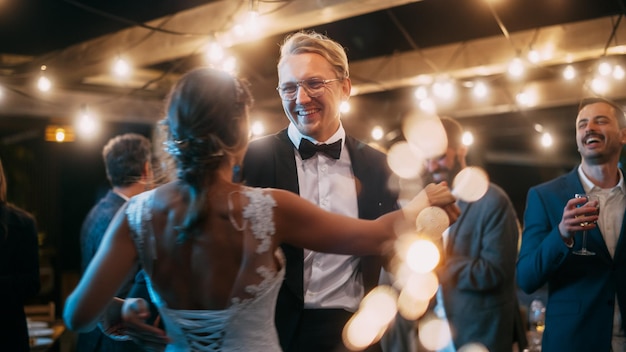 Mooie bruid in witte jurk en bruidegom in stijlvol zwart pak vieren bruiloft op een avondreceptie pasgetrouwden dansen op een locatie met beste multi-etnische diverse vrienden
