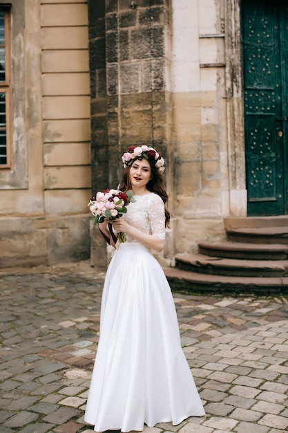 Mooie bruid met een boeket bloemen en een krans op de achtergrond van een oud gebouwx9