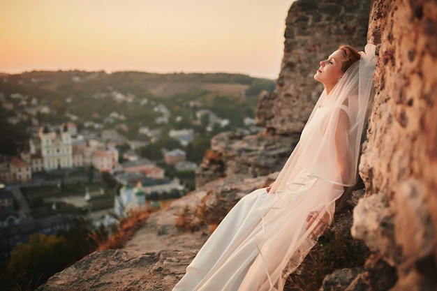 Mooie brunette bruid in een trouwjurk en een lange sluier poseren in de buurt van de bergen buiten de stad