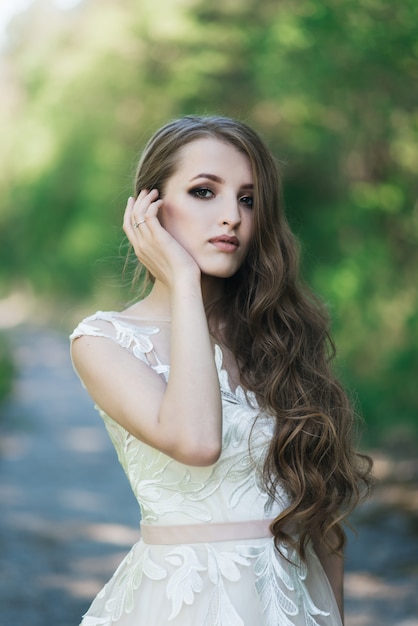 Mooie brunette bruid met krullen, make-up en stijlvolle jurk. portret op de muur van groene bomen. zomer trouwdag.