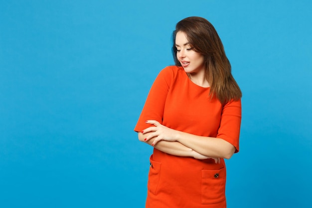 Mooie brunette jonge vrouw die een rode oranje jurk draagt en opzij kijkt staande poseren geïsoleerd over trendy blauwe muur achtergrond, studio portret. Mensen levensstijl mode concept. Bespotten kopie ruimte.