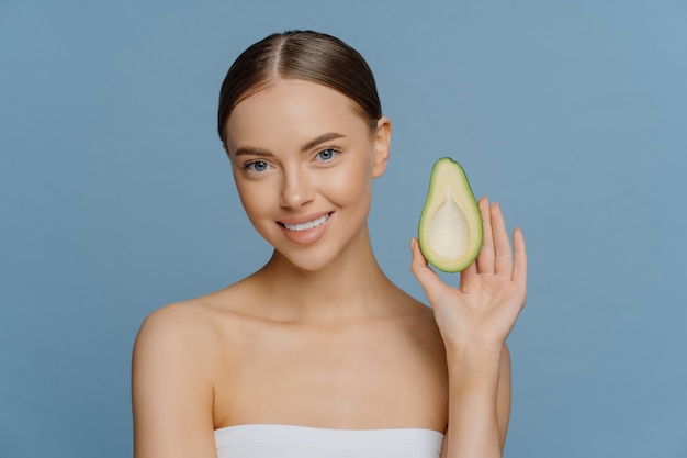 Mooie brunette jonge vrouw houdt de helft van avocado geniet van biologische cosmetica