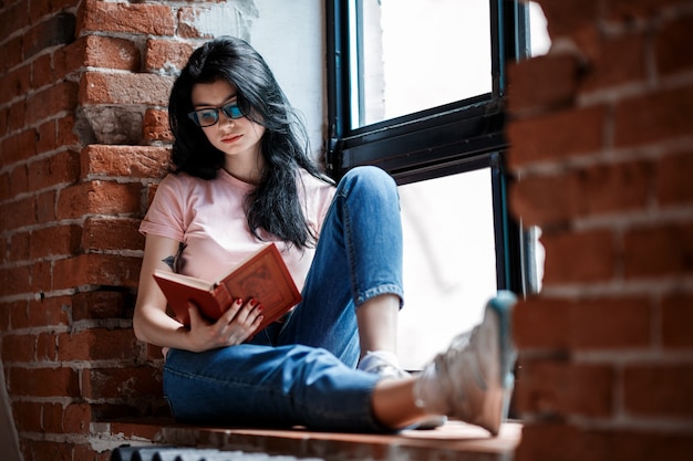 Mooie brunette jonge vrouw leesboek bij het raam thuis.