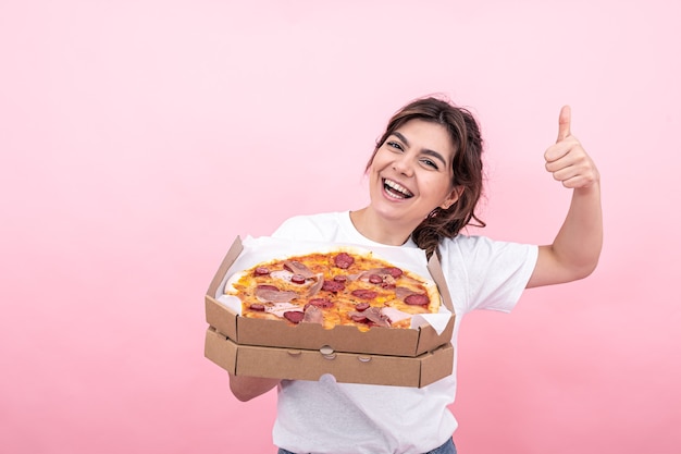 Mooie brunette meisje glimlachend met een doos pizza voor levering in haar hand roze achtergrond