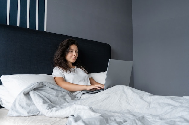 Mooie brunette meisje in bed met een laptop. Werk van 's ochtends tot' s avonds laat. Elektronische gadgets voor het slapengaan of 's ochtends.