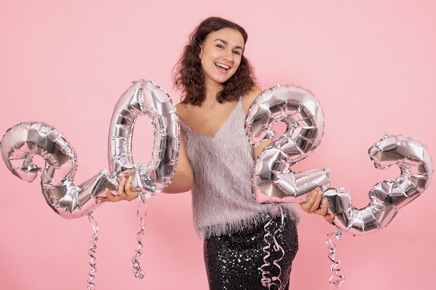 Mooie brunette meisje met krullend haar en feestelijke kleding poseren op een roze achtergrond met confetti en zilveren ballonnen uit de nummers 2023 te houden.