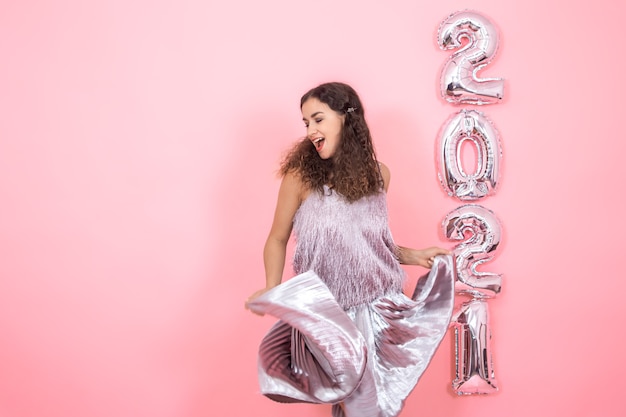 Mooie brunette meisje met krullend haar in feestelijke kleding in beweging op een roze muur met zilveren ballonnen voor het nieuwe jaar-concept