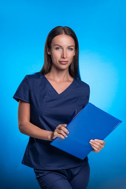 Mooie brunette met haar haren in een blauwe chirurgische pak poseren staande op een blauwe achtergrond, met een blauwe map en pen in haar handen. Verpleegster, glimlach, gezondheid.