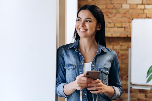 Mooie brunette vrouw die lacht met open mond en wegkijkt terwijl ze met een mobiele telefoon in een helder kantoor staat