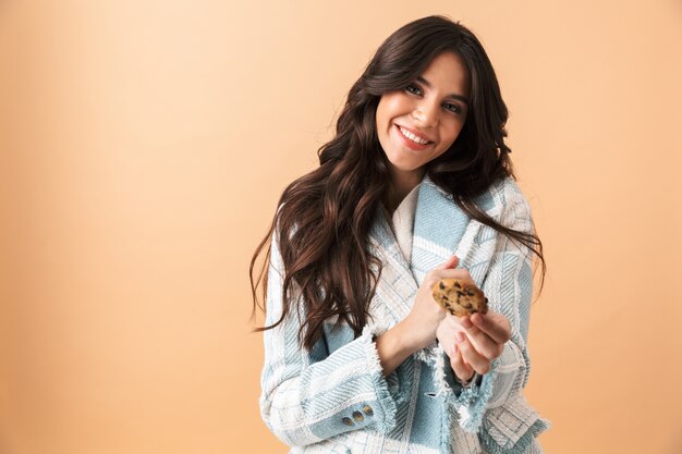 Mooie brunette vrouw gekleed in geruite jas staande geïsoleerd over beige, camera een koekje geven