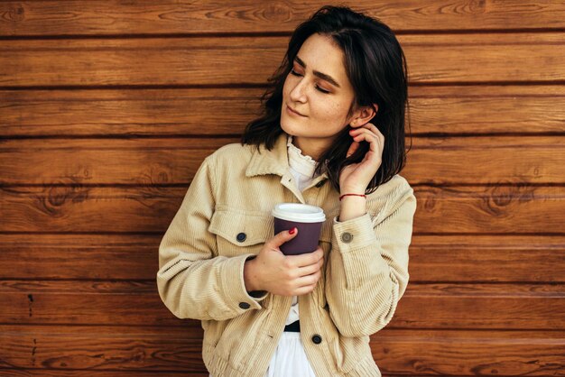 Mooie brunette vrouw houdt wegwerp kopje koffie in de straat van de stad Gelukkig vrouwelijke afhaalkoffie in handen Mooie student die 's ochtends buiten koffie drinkt terwijl ze naar de universiteit gaat