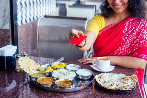 Mooie brunette vrouw in rode sari die met een plastic kaart betaalt voor traditionele thali met risecurddal en masala-thee