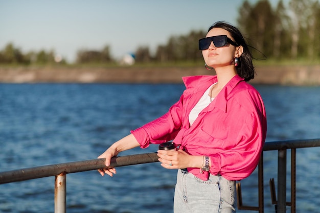 Mooie brunette vrouw in zonnebril met koffiekopje op pier aan de rand van het meer