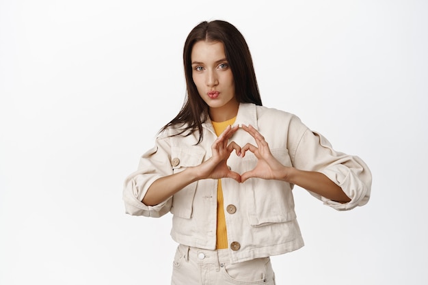 Mooie brunette vrouw tuit lippen zoenen, hart tonen ik hou van je teken, medeleven betuigen, haar gevoelens vertellen, zoals iets, staande in casual jas op wit