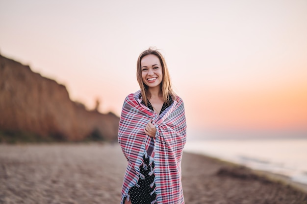 Mooie brunette vrouw wandelen op het strand bij zonsopgang gewikkeld in een warme deken.