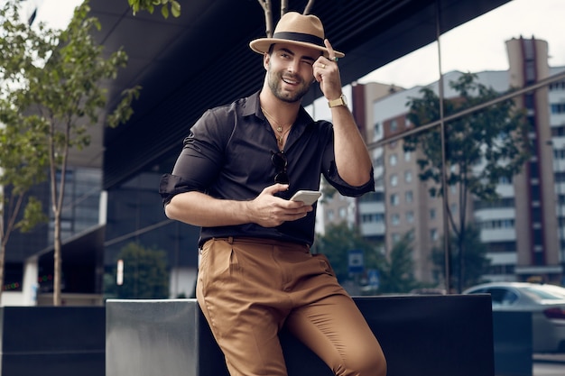 Mooie brutale gebruinde gespierde hipster man in zwart shirt en hoed met telefoon