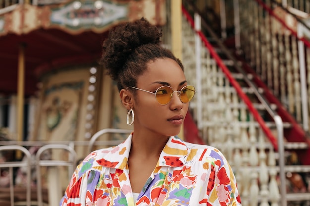 Mooie charmante krullende stijlvolle dame in kleurrijke, heldere zomerblouse en oranje zonnebril kijkt weg en poseert in de buurt van carrousel