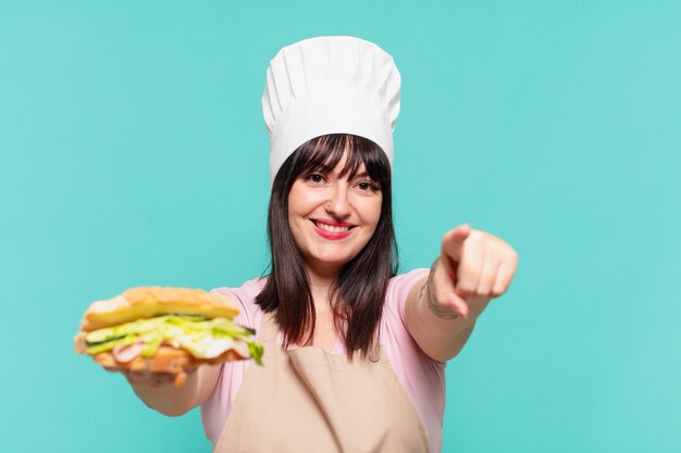 Mooie chef-kokvrouw die een sandwich wijst of toont en vasthoudt