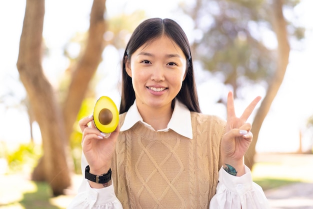 Mooie Chinese vrouw die een avocado vasthoudt in de open lucht glimlachend en overwinningsteken tonend