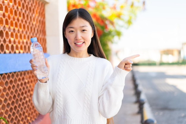 Mooie Chinese vrouw met een fles water in de buitenlucht verrast en wijzende vinger naar de zijkant