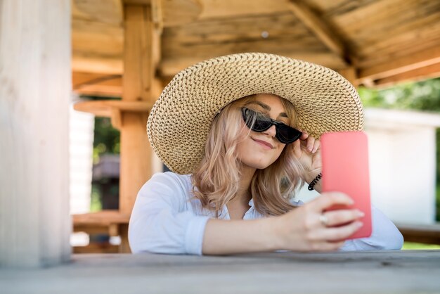 Mooie dame die cel vasthoudt en selfie doet in de buurt van houten prieel op hete zomerdag. ontspannen en genieten van de natuur