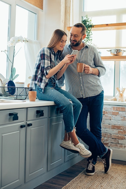 Mooie dame die in de keuken bij de gootsteen zit en smartphone gebruikt terwijl ze met vriendje is