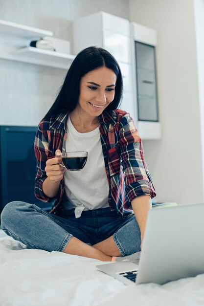 Mooie dame in jeans en shirt geniet van het werken in de laptop op haar bed terwijl ze koffie drinkt