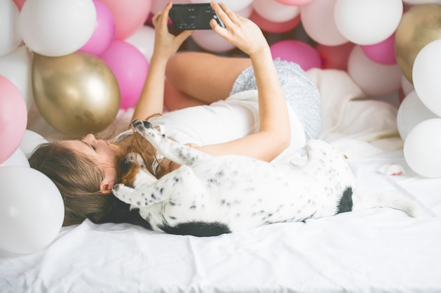 Mooie dame in pyjama selfie maken in haar slaapkamer met behulp van telefoon en rondhangen met haar hond.