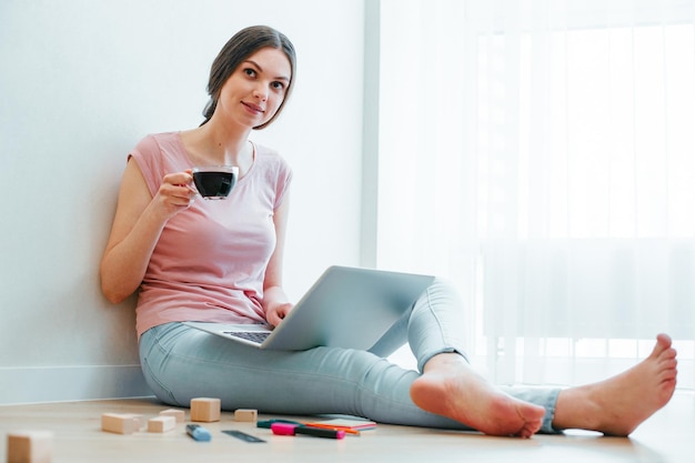 Mooie dame in vrijetijdskleding zittend op de vloer bij de muur met een laptop en een kopje en wegkijkend