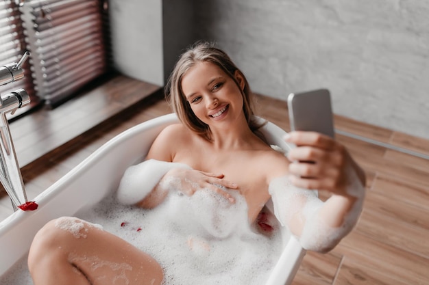 Mooie dame ontspannen in schuimend bad en selfie nemen op smartphone terwijl ze geniet van beauty morning